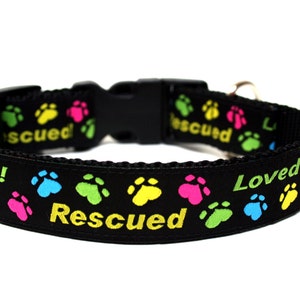 May include: Black dog collar with colorful paw prints and the words "Rescued! Loved!" in yellow and white. The collar has a black plastic buckle and a metal loop for attaching a leash. A pet accessory.