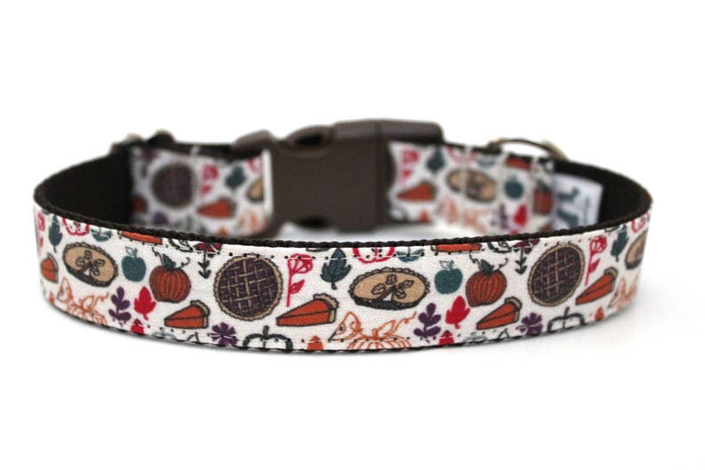 May include: A white dog collar with a brown adjustable buckle. The collar features a repeating pattern of fall-themed illustrations, including pumpkins, leaves, and pie slices.