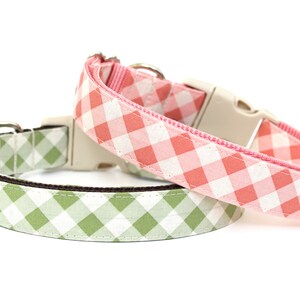 May include: Two dog collars with a gingham pattern. One collar is light green and white, the other is pink and white. Both collars have a beige plastic buckle and a silver metal ring.