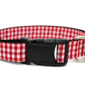 May include: Red and white gingham dog collar with a black plastic buckle and a silver metal D-ring. The collar is designed with a classic check pattern, suitable for pets.