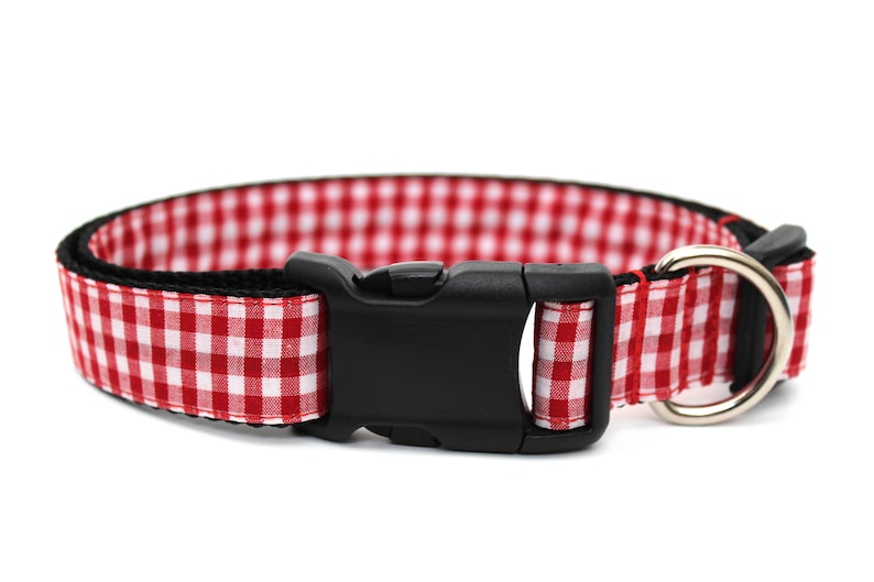 May include: Red and white gingham dog collar with a black plastic buckle and a silver metal D-ring. The collar is designed with a classic check pattern, suitable for pets.