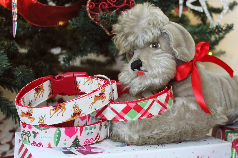 K&ouml;nnte beinhalten: Drei festliche Hundehalsb&auml;nder mit Weihnachtsmotiven. Die Halsb&auml;nder sind rot, wei&szlig; und gr&uuml;n mit Rentier-, Ornament- und Karomustern. Ein pl&uuml;schiger grauer Spielzeughund liegt auf den Halsb&auml;ndern.