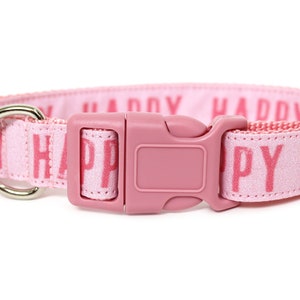 May include: Pink dog collar with a silver D-ring and a pink plastic buckle. The collar is made of a soft, woven fabric with the word "HAPPY" printed repeatedly in pink.