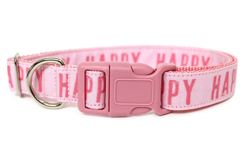 May include: Pink dog collar with a silver D-ring and a pink plastic buckle. The collar is made of a soft, woven fabric with the word "HAPPY" printed repeatedly in pink.