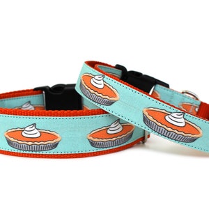 May include: Two dog collars with a turquoise background and a pattern of orange pumpkin pies with white whipped cream on top. The collars are red on the inside and have black buckles.