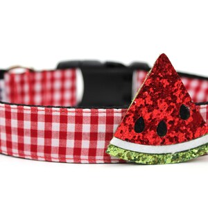 May include: A red and white gingham dog collar with a black buckle and a watermelon-shaped embellishment. The watermelon slice is red with black seeds and a green glitter rind.