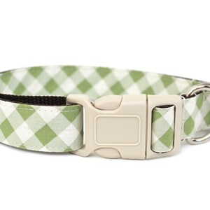 May include: A dog collar with a green and white gingham pattern. The collar has a beige plastic buckle and a silver metal D-ring for leash attachment. The interior of the collar is a dark brown color.