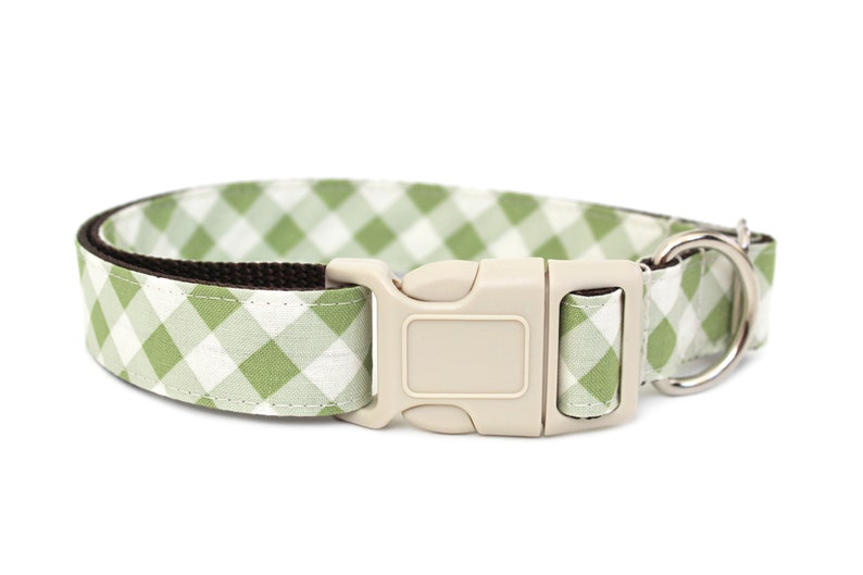 May include: A dog collar with a green and white gingham pattern. The collar has a beige plastic buckle and a silver metal D-ring for leash attachment. The interior of the collar is a dark brown color.