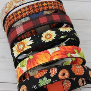 May include: A stack of colorful dog collars with various autumnal patterns, including sunflowers, pumpkins, and leaves. The collars are made of fabric and have a black buckle.