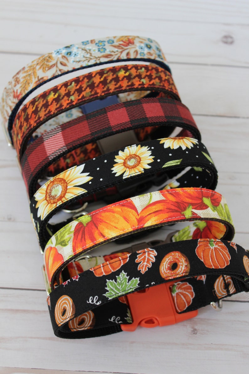 May include: A stack of colorful dog collars with various autumnal patterns, including sunflowers, pumpkins, and leaves. The collars are made of fabric and have a black buckle.