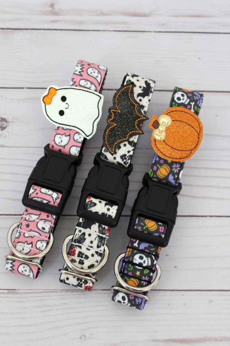 May include: Three Halloween-themed dog collars. One is pink with ghost patterns and a ghost applique. The second is black and white with a bat applique. The third is purple with pumpkin and skull patterns and a pumpkin applique. Each has a black buckle.