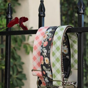 May include: Three dog collars hang on a black metal fence. One is pink gingham, one is black floral, and the third is green gingham. The collars have silver metal hardware and are displayed outdoors.