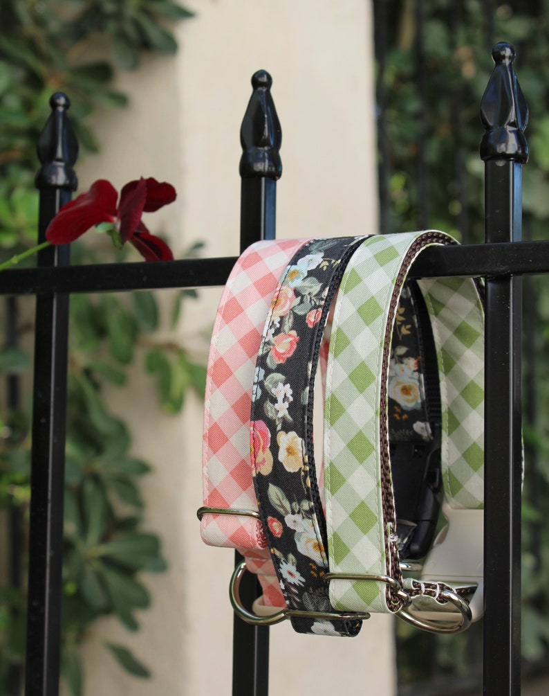 May include: Three dog collars hang on a black metal fence. One is pink gingham, one is black floral, and the third is green gingham. The collars have silver metal hardware and are displayed outdoors.