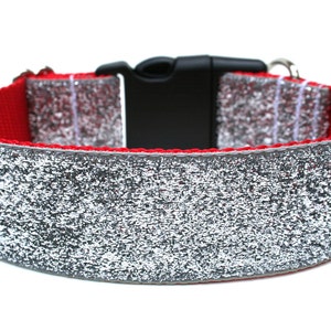 May include: A dog collar with a red interior and a silver glitter exterior. The collar has a black plastic buckle and a silver metal loop for attaching a leash. The collar is designed for pets.