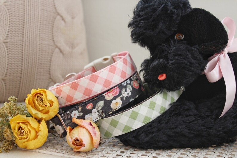 May include: Three dog collars with different patterns: pink gingham, floral, and green gingham. A black stuffed dog wears one of the collars. Dried yellow roses and a beige pillow are in the background.