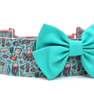 May include: A turquoise blue dog collar with a matching bow tie. The collar is printed with a pattern of sea creatures, including fish, jellyfish, seahorses, and an octopus.