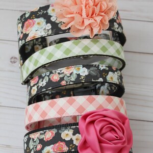 May include: A stack of dog collars with floral and gingham patterns. The collars feature a variety of colors, including black, pink, and green. Each collar is adorned with a decorative flower in shades of pink.