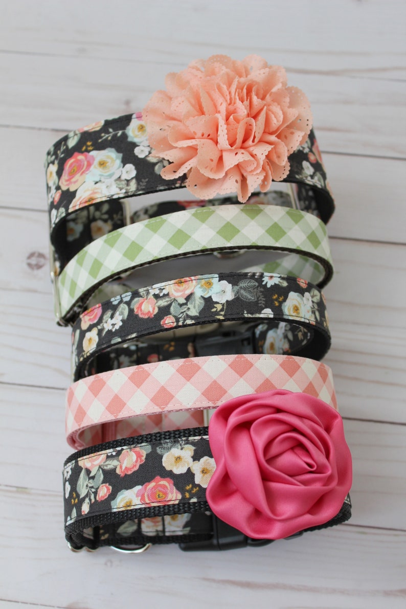 May include: A stack of dog collars with floral and gingham patterns. The collars feature a variety of colors, including black, pink, and green. Each collar is adorned with a decorative flower in shades of pink.