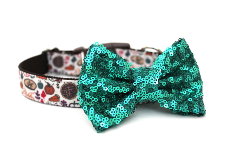 May include: A green sequin bow tie attached to a white and brown dog collar with a Thanksgiving-themed print featuring pumpkins, pies, and leaves.