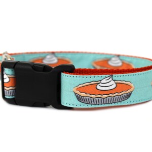 May include: A turquoise blue dog collar with a red trim and a repeating pattern of pumpkin pies with whipped cream. The collar has a black plastic buckle.