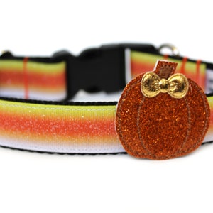 May include: A Halloween-themed dog collar with a candy corn pattern in yellow, orange, and white. The collar has a black buckle and a sparkly orange pumpkin charm with a gold bow.