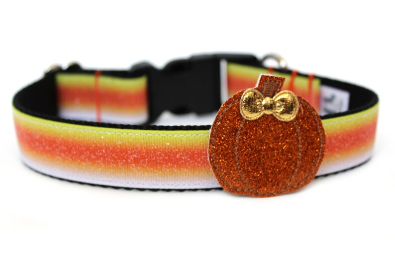 May include: A Halloween-themed dog collar with a candy corn pattern in yellow, orange, and white. The collar has a black buckle and a sparkly orange pumpkin charm with a gold bow.