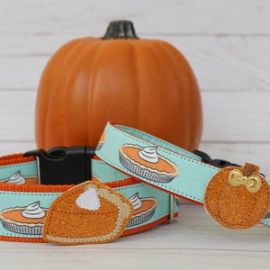May include: Dog collar set with a pumpkin pie pattern on a light blue background. The set includes a collar, a charm, and a pumpkin-shaped charm with a gold bow. An orange pumpkin is in the background.