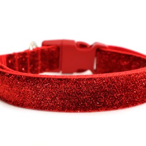 Könnte beinhalten: Ein leuchtend rotes Haustierhalsband mit einer dichten, glitzernden Oberfläche auf der gesamten Außenseite. Das Halsband verfügt über eine passende rote Kunststoff-Steckschnalle, einen metallenen D-Ring zur Befestigung und ein verstellbares Schiebeelement.