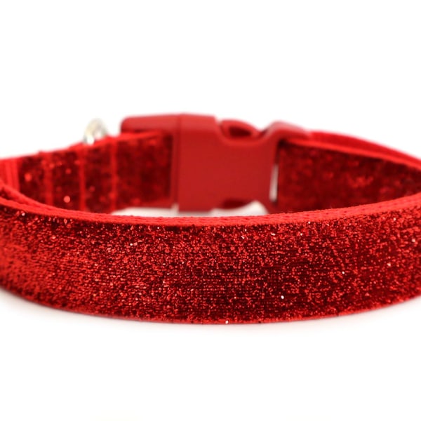 May include: A bright red pet collar featuring a dense, sparkling glitter finish across the entire outer surface. The collar includes a matching red plastic side-release buckle, a metal D-ring for attachment, and an adjustable sliding hardware component.