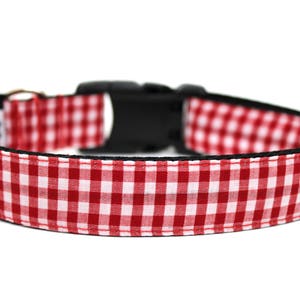 May include: A red and white gingham dog collar with a black plastic buckle. The collar is made of fabric with a classic check pattern. The collar is designed for pets.