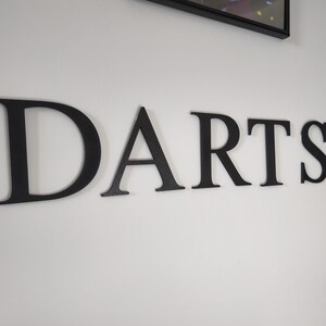 Könnte beinhalten: Schwarze Holzbuchstaben mit der Aufschrift "DARTS" sind an einer weißen Wand angebracht. Die Buchstaben sind in einer Serifenschrift und horizontal angeordnet. Rechts vom Wort befindet sich eine schwarze Steckdose. Ein gerahmtes Bild hängt über dem Wort.