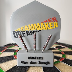 Könnte beinhalten: Eine Dartscheibe mit einem individuellen Dart-Halter, der drei Darts hält. Der Halter trägt den Text "DREAMMAKER" und "Dimitri Van den Bergh". Die Darts sind silberfarben mit roten Akzenten. Das Dartboard ist schwarz, weiß und beige.