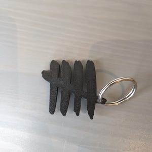 May include: Black metal keychain featuring a tally mark design. The keychain has a silver-colored split ring for attaching keys. The tally mark is a series of five vertical lines, with the fifth line crossing the first four.