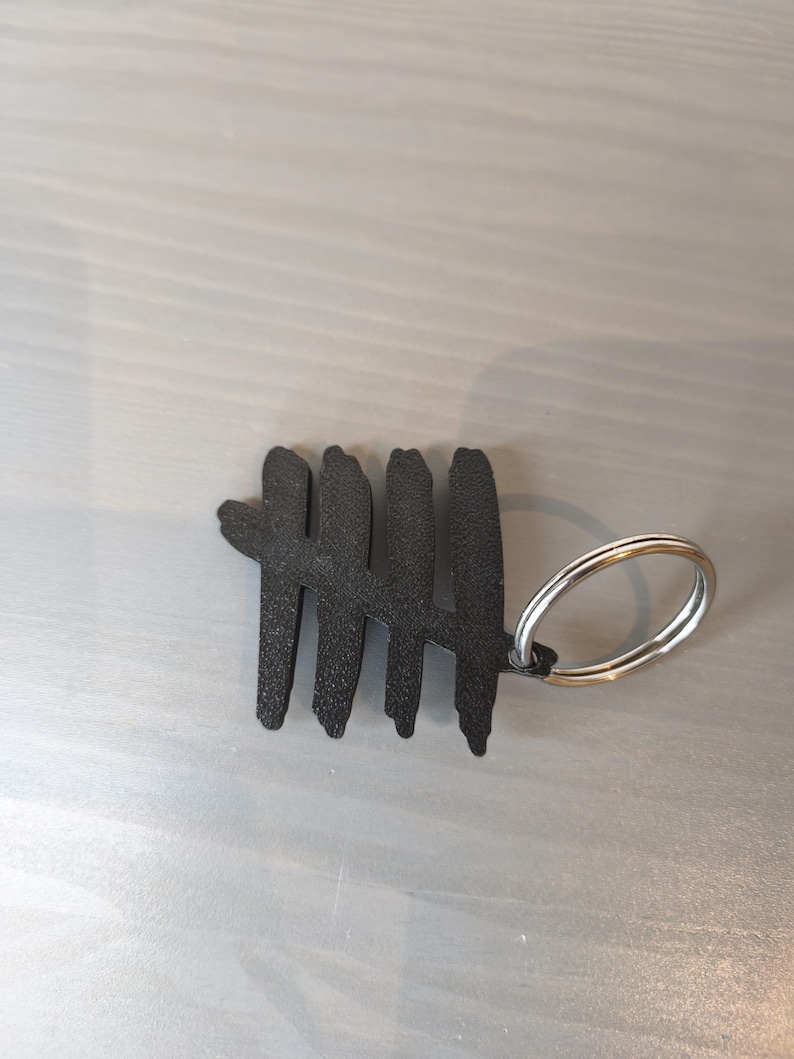 May include: Black metal keychain featuring a tally mark design. The keychain has a silver-colored split ring for attaching keys. The tally mark is a series of five vertical lines, with the fifth line crossing the first four.