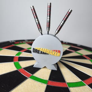 Könnte beinhalten: Ein grauer Dart-Halter mit dem Wort "DREAMMAKER" in Schwarz, Rot und Gelb. Drei Darts mit silbernen und roten Details sind in den Halter eingesetzt, platziert auf einer Dartscheibe mit schwarzen, grünen, roten und beigen Abschnitten.