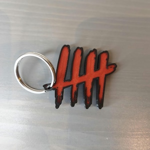 May include: A red and black keychain featuring a stylized design of five vertical lines. The lines have a distressed, textured appearance. The keychain includes a silver-colored metal ring for attaching keys.