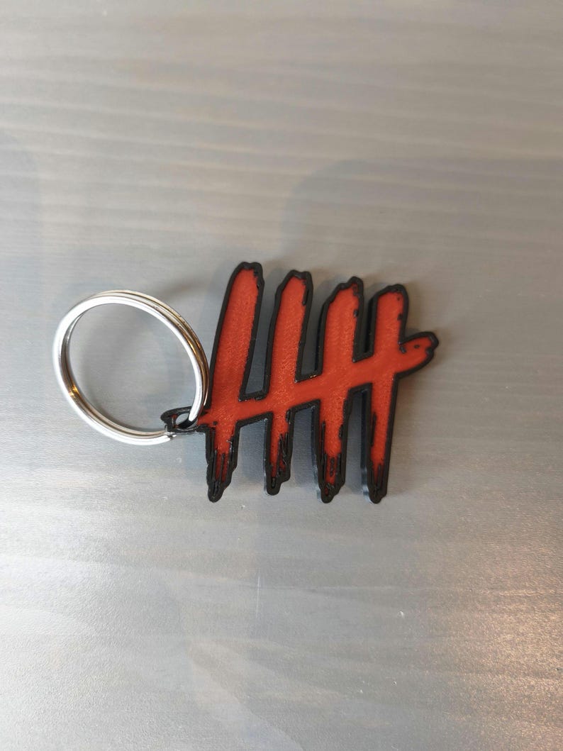 May include: A red and black keychain featuring a stylized design of five vertical lines. The lines have a distressed, textured appearance. The keychain includes a silver-colored metal ring for attaching keys.