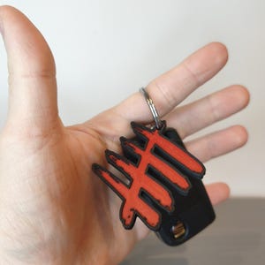May include: A black and red keychain with a unique design. The keychain features a red symbol on a black base, attached to a silver key ring. The design is held in a hand against a neutral background.