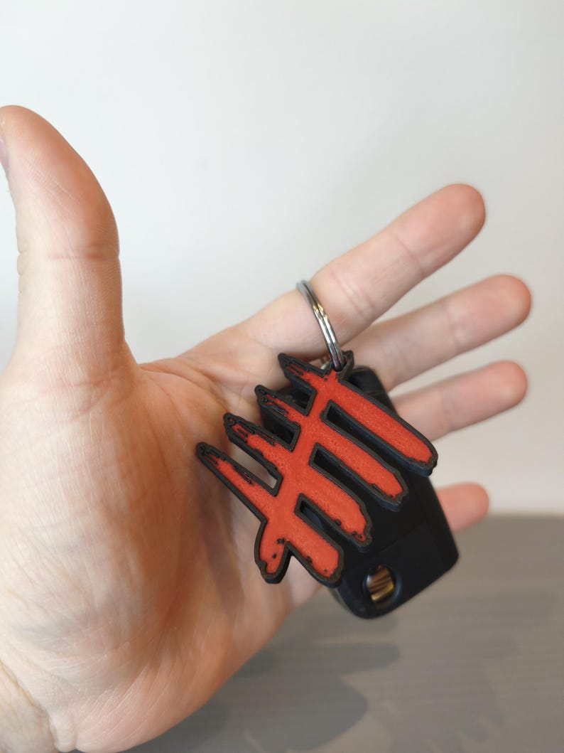 May include: A black and red keychain with a unique design. The keychain features a red symbol on a black base, attached to a silver key ring. The design is held in a hand against a neutral background.