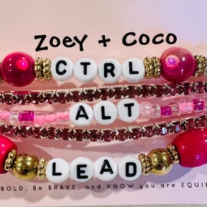 May include: Stacked beaded bracelets with the words "CTRL", "ALT", and "LEAD" spelled out in white letter beads. The bracelets feature pink, gold, and white beads. The text "Zoey + Coco" and "Be BOLD. Be BRAVE. and KNOW you are EQUIPPED" are also visible.