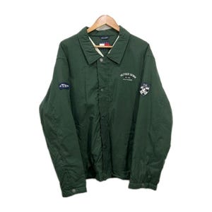 May include: A dark green jacket with a button-up front and a collar. The jacket has the text "HILFIGER DENIM" and "TH" patches on the sleeves. The jacket is hanging on a wooden hanger.