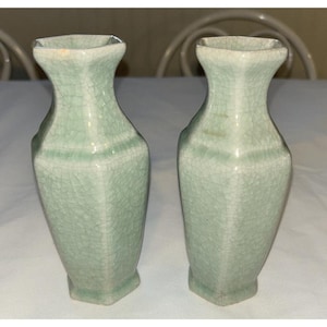 May include: Two celadon-colored ceramic vases with a crackle glaze. The vases have a geometric shape with a square base and a flared, square top. The vases are displayed on a white surface.