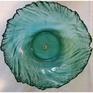 Op de afbeelding: Een teal glazen kom met een gerimpelde rand. De kom heeft een cirkelvormige basis en een getextureerd, wervelend patroon. De randen zijn ongelijkmatig, wat bijdraagt aan het unieke ontwerp van de kom. Een klein label is zichtbaar in het midden.