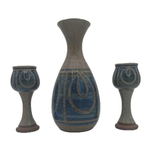 May include: A ceramic set consisting of one tall, narrow-necked carafe and two matching stemmed goblets. Each piece features a speckled light gray base with horizontal blue bands and a repeating hand-painted blue swirl pattern around the body.