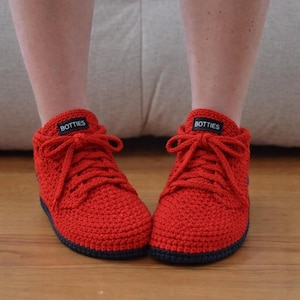 May include: Red crochet sneakers with black soles and laces. Each shoe has the word "BOTTIES" on the tongue. These handmade sneakers are a cosy and stylish footwear option.