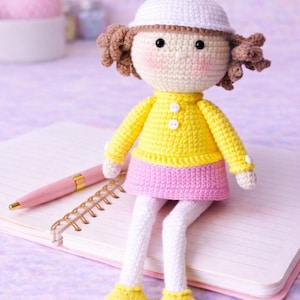 May include: A handmade crochet doll with a white bunny ear hat, brown curly hair, and rosy cheeks. The doll wears a yellow sweater, pink skirt, white leggings, and yellow shoes. It sits on a notebook with a pink pen.
