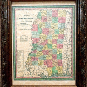 May include: A vintage map of Mississippi with its roads and distances. The map is framed in a dark brown wooden frame with ornate details. The map is printed on paper with a cream background and features a green border.