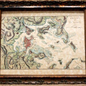 May include: Framed antique map of Boston and its environs, featuring detailed cartography with hand-colored elements. The map is encased in a dark, ornate wooden frame, adding a vintage aesthetic. The text "BOSTON et SES ENVIRONS" is visible.