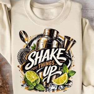 Könnte beinhalten: Cremefarbenes Sweatshirt mit einem Cocktail-Motiv. Das Design umfasst einen Cocktailshaker, einen Jigger, einen Löffel und den Schriftzug "Shake Things Up" in einer kräftigen, stilisierten Schriftart. Frische Limette, Minze und Eiswürfel vervollständigen das Design.