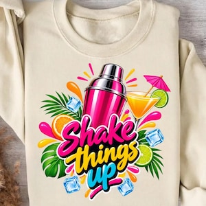 May include: A beige sweatshirt featuring a graphic of a pink cocktail shaker, a glass of orange drink with a pink umbrella, lime and orange slices, ice cubes, and green palm leaves. The text reads Shake things up in pink and yellow.